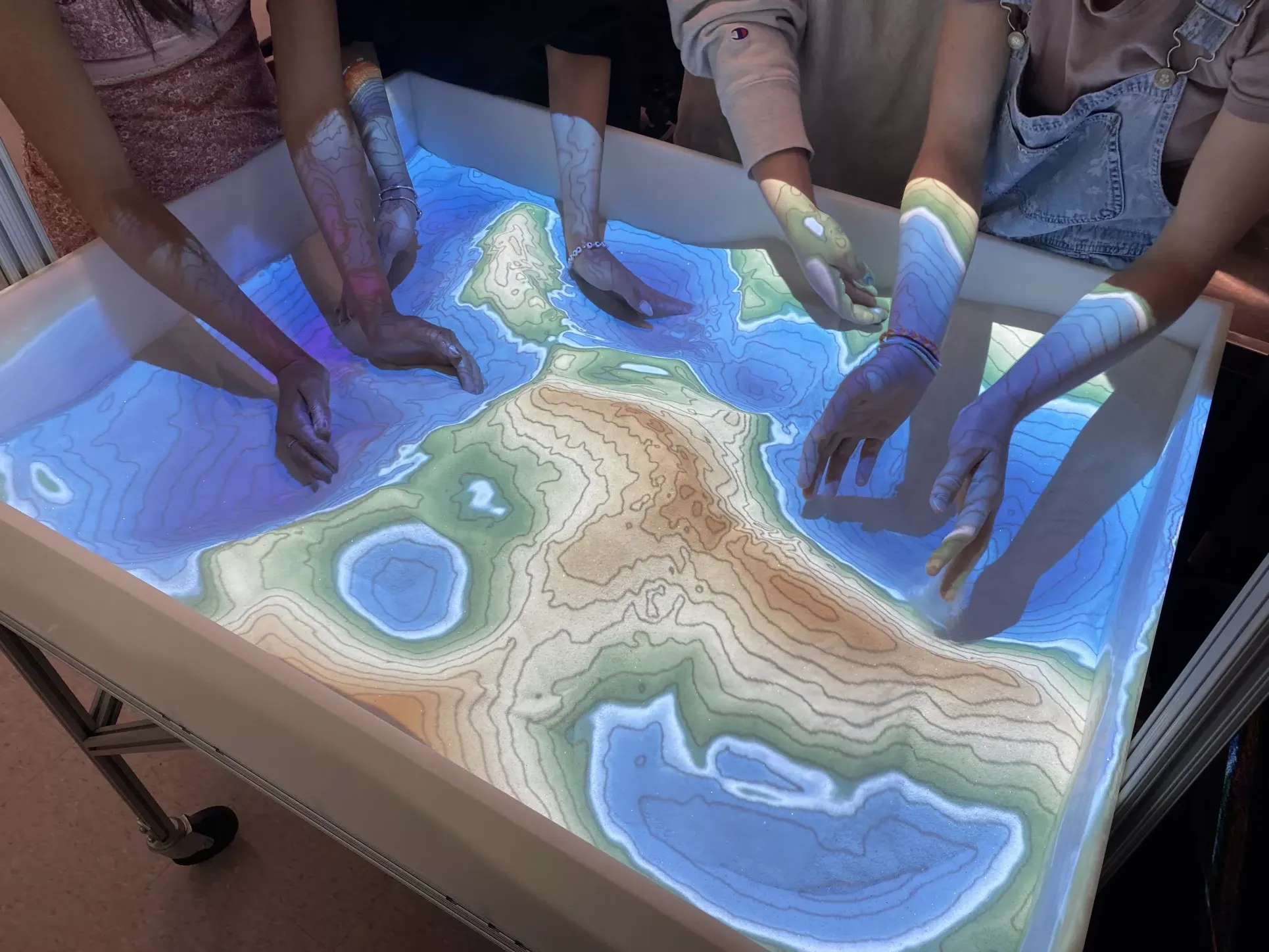 Several students put their hands into the sandbox. The digital map projected on the sand changes as the students move the sand around.